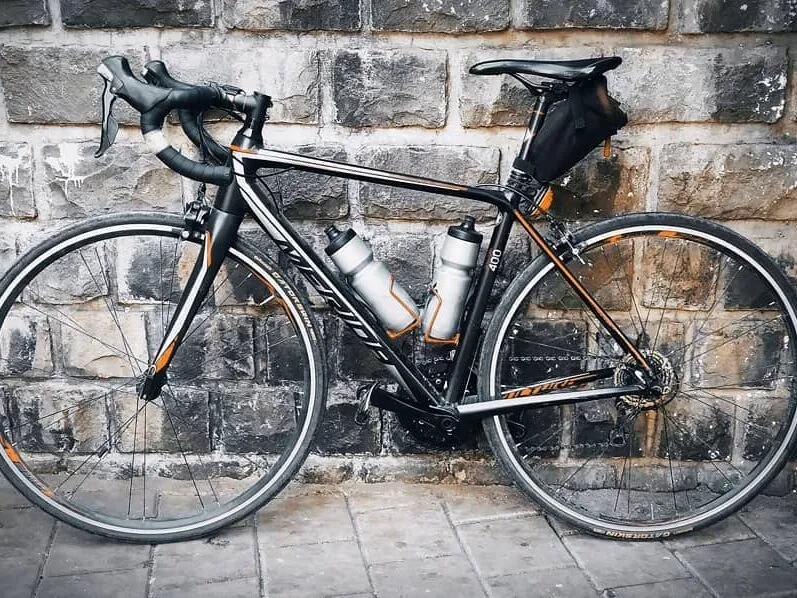 A black road bike with two bottles and a saddle bag, leaning against a stone wall.